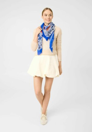 Junge Frau trägt beigen Pullover, weißen Rock, weiße Sneaker und blauen gemusterten Schal, steht lächelnd vor weißem Hintergrund.