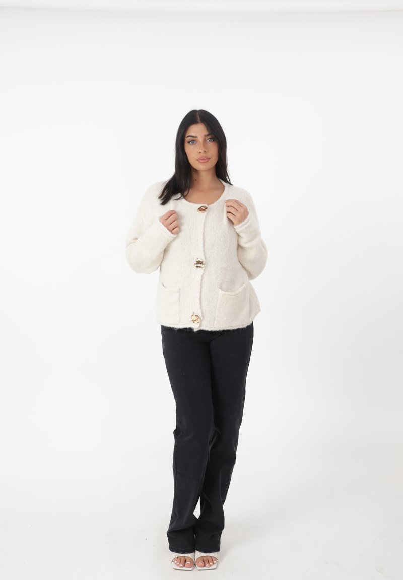 Cardigan in maglia color avorio con scollo rotondo, tre grossi bottoni, due tasche frontali e polsini a coste, abbinato a pantaloni neri aderenti.