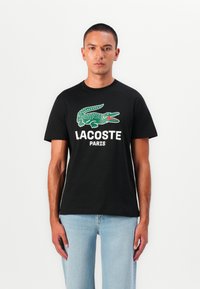 Sort bomulds t-shirt med et grønt krokodille-logo og teksten "LACOSTE PARIS" i hvid. Fit'et er afslappet med korte ærmer.