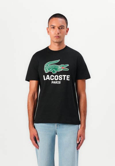 Черна памучна тениска с зелен логотип на крокодил и текст "LACOSTE PARIS" в бяло. Фитът е свободен с къси ръкави.