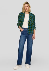 Grüner und blauer Blazer mit Hahnentrittmuster, weißes T-Shirt, hoch taillierte blaue Jeans und weiße Turnschuhe, stehend vor einem grauen Hintergrund.