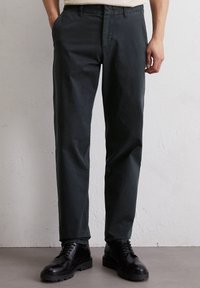 Pantalons en coton vert foncé avec une coupe droite, présentant un design classique à cinq poches et une fermeture à bouton. Stylés avec des bottes à lacets noires.