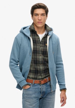 Felpa blu con zip e cappuccio regolabile sopra una camicia a quadri nera e verde. L'outfit include jeans azzurri chiari e una cintura marrone.