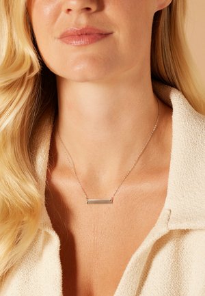 Zilveren barenketting op een dunne ketting, rustend op gladde huid; heeft een minimalistisch ontwerp en een gepolijste afwerking. Crème-kleurige stof achtergrond.