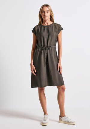 Robe vert olive à manches courtes avec une taille cintrée à cordon. Tissu doux et lisse avec une silhouette mi-genoux. Portée avec des baskets blanches.