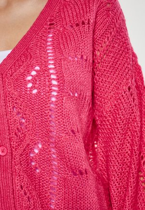 Cardigan en tricot rose avec un décolleté en V, des motifs complexes et une fermeture à boutons. La texture est légère avec des sections à mailles ajourées.