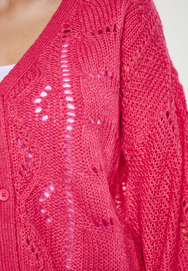 Vaaleanpunainen neulecardigan, jossa on V-pääntie, monimutkaisia kuvioita ja nappikiinnitys. Kankaan rakenne on kevyt ja siinä on avoimia kudontakohtia.