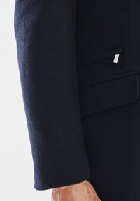 Morska wełniana płaszcz z gładką fakturą, widocznymi szwami i subtelnym detalem kieszeni. Posiada małą metkę z logo w pobliżu kieszeni.