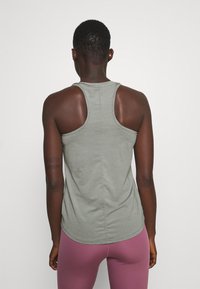 Canotta atletica verde chiaro con design a schiena scoperta, texture morbida e orlo arrotondato. Indossata con leggings viola aderenti.