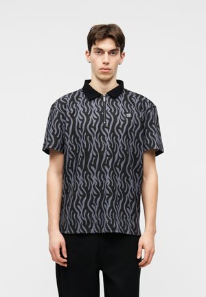 Ung mand iført en sort og grå mønstret poloshirt med en kort lynlås og sort krave, stående foran en ensfarvet lys baggrund.