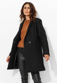 Manteau en laine noir à double boutons, avec des revers, et des poches latérales, associé à un pull marron et un pantalon en cuir noir.