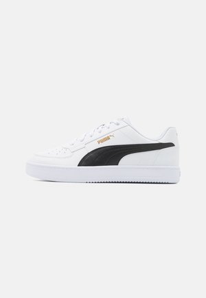 Hvid sneaker med sort PUMA-logo, har en glat læderoverdel, strukturerede overlays, rund tå og polstret krave, understøttet af en gummisål.