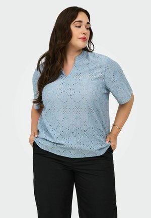 Kvinne med langt mørkt hår iført en lyseblå mønstret kortermet bluse og svarte bukser, hender i lommer, ser litt til siden.