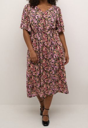 Kvinde iført en knælængde blomsterkjole i pink, sort og beige, med korte flagrende ærmer og sorte stroppesandaler, stående mod en ensfarvet væg.