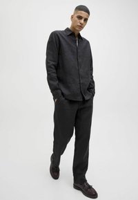Chemise noire en lin à manches longues avec un col boutonné, associée à un pantalon noir en lin. Les chaussures sont en cuir brun foncé avec des lacets décoratifs.