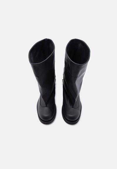 Ζευγάρι ψηλές μαύρες δερμάτινες μπότες που στέκονται όρθιες σε απλό λευκό φόντο, οπτική από πάνω.