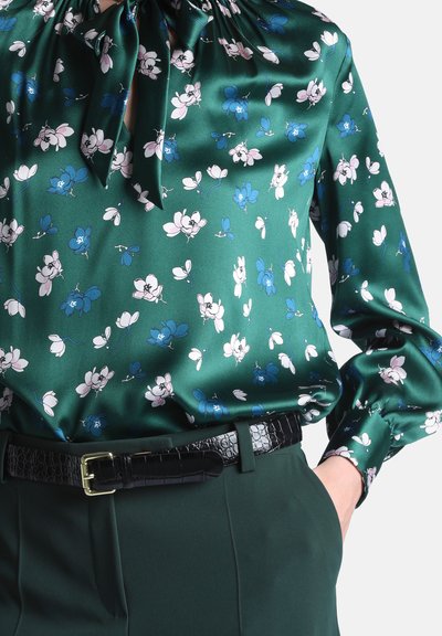 Blouse en satin verte avec imprimé floral bleu et blanc, noeud au niveau du col, associée à un pantalon vert foncé et une ceinture noire texturée.