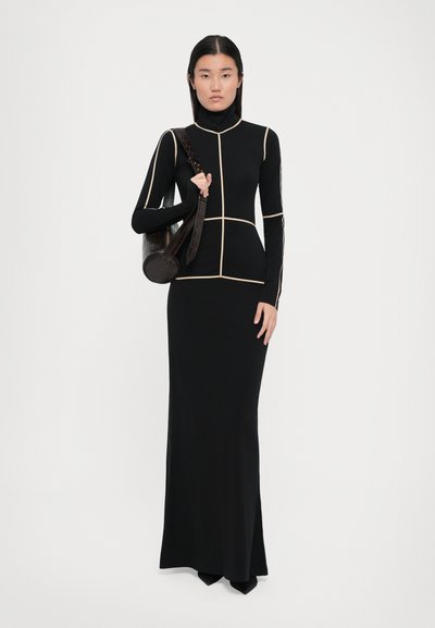 Black long-sleeve dress with a high neckline, featuring a fitted design and beige geometric piping. Model holds a round, brown handbag.