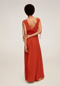 Femme à cheveux courts et bouclés portant une longue robe rouge sans manches avec un dos en V profond, se tenant avec un bras croisé derrière elle.