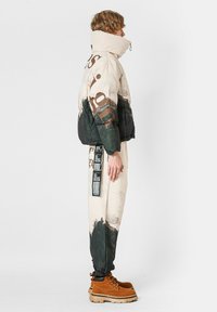 Chaqueta acolchada con un esquema de color beige y negro, con un estampado de naturaleza y letras prominentes. Combinada con pantalones que muestran un patrón de bosque.