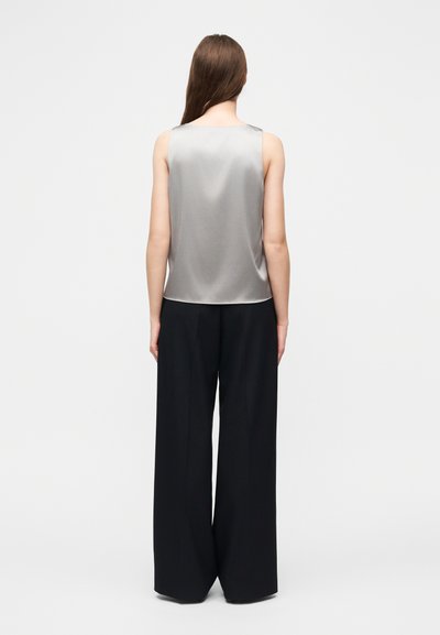 Femme aux longs cheveux bruns portant un haut argenté sans manches et un pantalon noir large et ample, debout de dos sur un fond blanc uni.