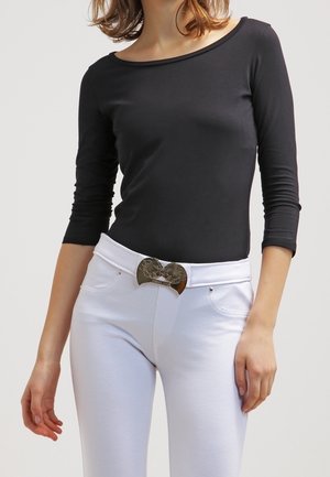 Frau trägt ein schwarzes langärmeliges Oberteil und hoch taillierte weiße Hose mit dekorativer Metallschnalle am Gürtel.