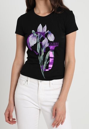 Nainen, joka pukeutuu mustaan istuvaan t-paitaan, jossa on violetti "GUESS"-logo ja valkoinen kukkakuosi, yhdistettynä valkoisiin housuihin, yksinkertaisen taustan edessä.