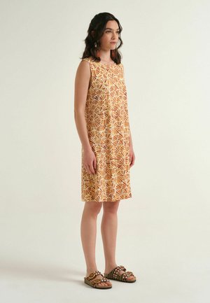 Donna in piedi con un abito senza maniche arancione e bianco a motivo, lungo fino al ginocchio, con sandali aperti su uno sfondo semplice.