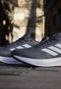 Paire de chaussures de running Adidas grises avec des bandes blanches et des semelles intermédiaires Cloudfoam posées sur un pavé en pierre à l'extérieur.