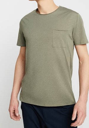 Mand iført en olivengrøn t-shirt med korte ærmer og brystlomme, kombineret med mørkeblå bukser, mod en ensfarvet baggrund.