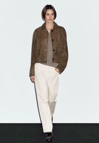Model pewnie maszeruje, nosząc brązową liderkę, teksturowany sweter i białe spodnie, na minimalistycznym tle.