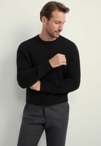 Czarny sweter dzianinowy z okrągłym dekoltem, dopasowanymi mankietami i długimi rękawami, w połączeniu z szarymi spodniami. Gładka faktura z minimalnymi detalami.