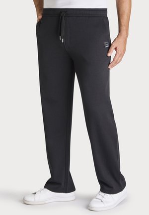 Mann trägt schwarze Jogginghose mit Kordelzugbund und weiße Turnschuhe, steht mit einer Hand in der Tasche und der anderen locker herunterhängend.