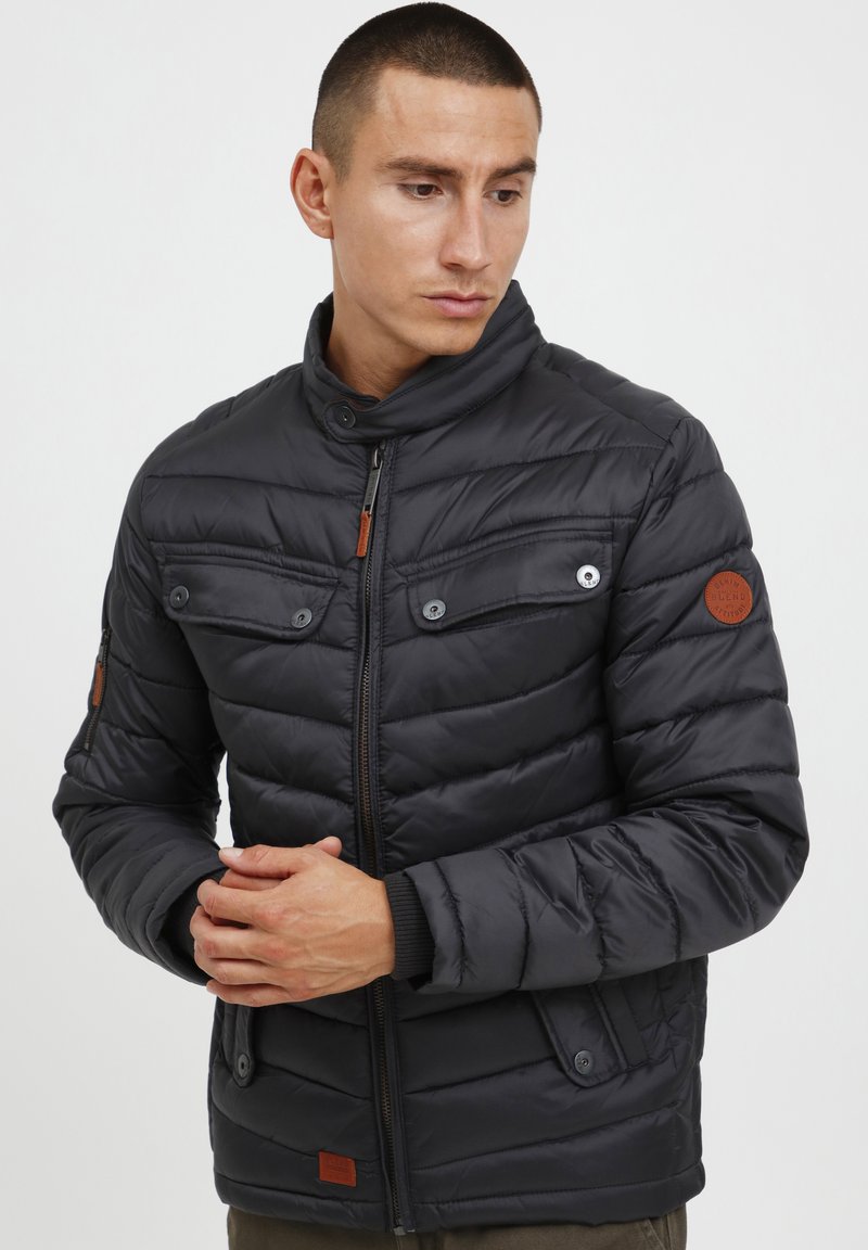Veste matelassée noire avec un col haut, dotée de poches à bouton et d'un accent en cuir sur la manche. Texture lisse avec un design épuré.