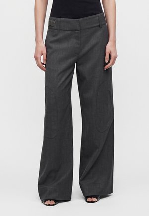 Femme portant un pantalon large gris foncé avec des renforts subtils aux genoux et des talons noirs à bouts ouverts, debout devant un fond uni.