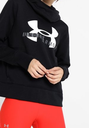Osoba ubrana w czarną bluzę z kapturem Under Armour z dużym logo oraz czerwone legginsy z małym logo widocznym na udzie na białym tle.