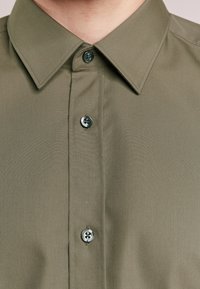 Camisa verde oliva con cuello puntiagudo, con cierre frontal de botones y botones grises a contraste, confeccionada en tejido liso.