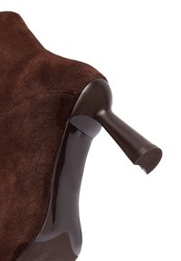 Scarpa con tacco alto in suede marrone, con tacco in plastica lucida marrone scuro. Presenta una punta arrotondata e una superficie liscia con una sottile texture.