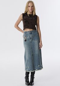 Braunes, kurzes Oberteil mit Rüschen, auf dem der Text "Made of" steht. Hellblaue Denim-Maxirock mit fransigem Saum und dekorativem Kettendetail. Schwarze Stiefeletten.