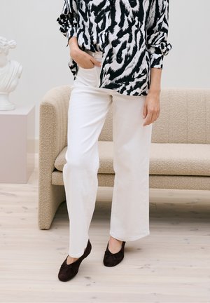 Personne portant un pantalon blanc à jambes larges, une chemise à manches longues noire et blanche à motifs, et des ballerines noires, se tenant près d'un canapé beige et d'une sculpture de buste.