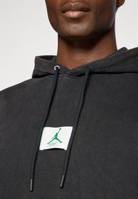 Sort hættetrøje med runde snore og et frontlogo-patch med en grøn silhuet. Stof med en tekstureret overflade.