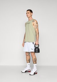 Light green sleeveless top with "HYROX" logo, white shorts, black kettlebell, and black/red athletic shoes. Tattoos visible on arms and legs.