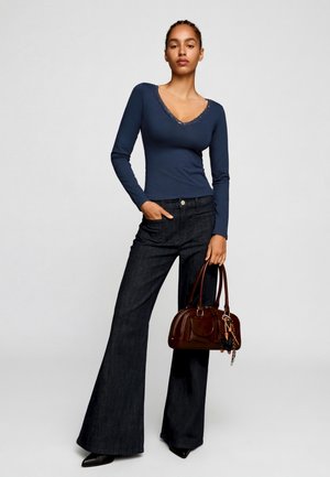 Ženska, oblečena v temno modro majico z dolgimi rokavi in V-izrezom ter širokimi temno modrimi kavbojkami, drži rjavo torbico s obeskom za ključe.