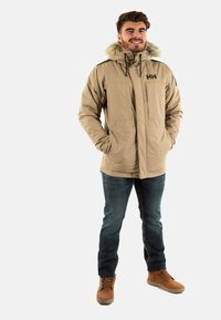 Giacca beige isolante con cappuccio foderato in pelliccia, chiusura con zip e tasche laterali, abbinata a jeans blu e stivali marroni.