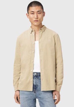 Ung mand iført beige skjorte med knapper over en hvid tanktop og blå jeans, stående mod en ensfarvet lys baggrund.