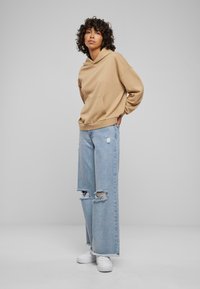 Sudadera beige con un bolsillo frontal, combinada con jeans de pierna ancha en azul claro que presentan dobladillos desflecados y detalles desgastados. Zapatillas blancas completan el conjunto.