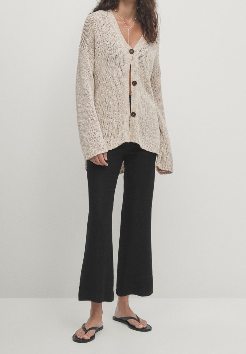 Femme portant un cardigan en maille beige oversize, un pantalon noir à jambes larges, des tongs noires et un simple collier argenté sur fond uni.