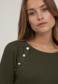 Pulover ribbed de culoare verde măslinie, cu nasturi argintii pe linia umărului, cu guler rotund și textură netedă.