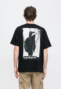 Czarna bawełniana koszulka z dużą monochromatyczną grafiką na plecach przedstawiającą postać w kapturze; u dołu napis "carhartt".