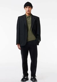 Blazer negro oversized sobre una camiseta polo verde olivo, combinado con pantalones negros y zapatillas negras, con un gorro de canalé y acentos en contraste.
